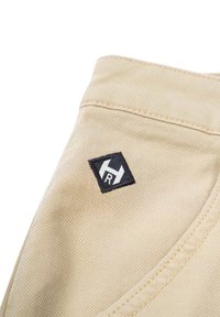 Beige Stoff mit einer strukturierten Oberfläche und sichtbaren Nähten. Verfügt über ein schwarzes, rautenförmiges Logopatch mit einem weißen Emblem.
