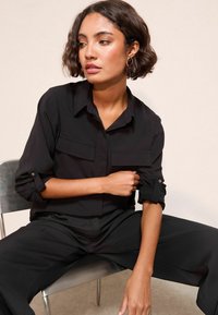 Camicia nera con bottoni e maniche arrotolabili, dotata di due tasche sul petto, realizzata in un tessuto liscio e leggero. Si abbina a pantaloni neri larghi.