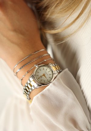 Zilveren en gouden roestvrijstalen horloge met een witte wijzerplaat, gouden uuraanduiders en datumfunctie, gedragen met meerdere zilveren armbanden.