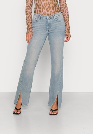 Jeans de ganga azuis claros com boca de sino e fendas laterais na bainha. Com um estilo clássico de cinco bolsos e um ajuste de altura média. Usados com chinelos pretos.