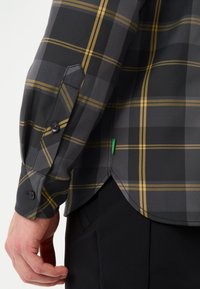 Camicia a quadri nera e grigia con accenti gialli, dotata di polsini con bottone e orlo curvo. Il tessuto sembra morbido e leggero.