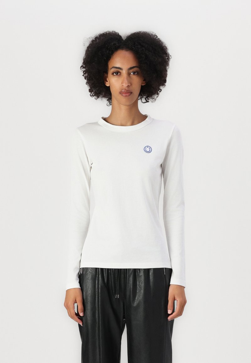 T-shirt bianco a maniche lunghe realizzato in tessuto morbido, con un piccolo logo di faccina sorridente blu sul lato sinistro del petto. Collo rotondo e taglio aderente.