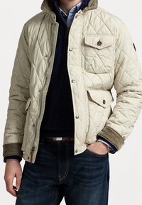 Quilted cream jacket with a corduroy collar, snap button pockets, and ribbed cuffs, worn over a dark sweater and denim jeans.