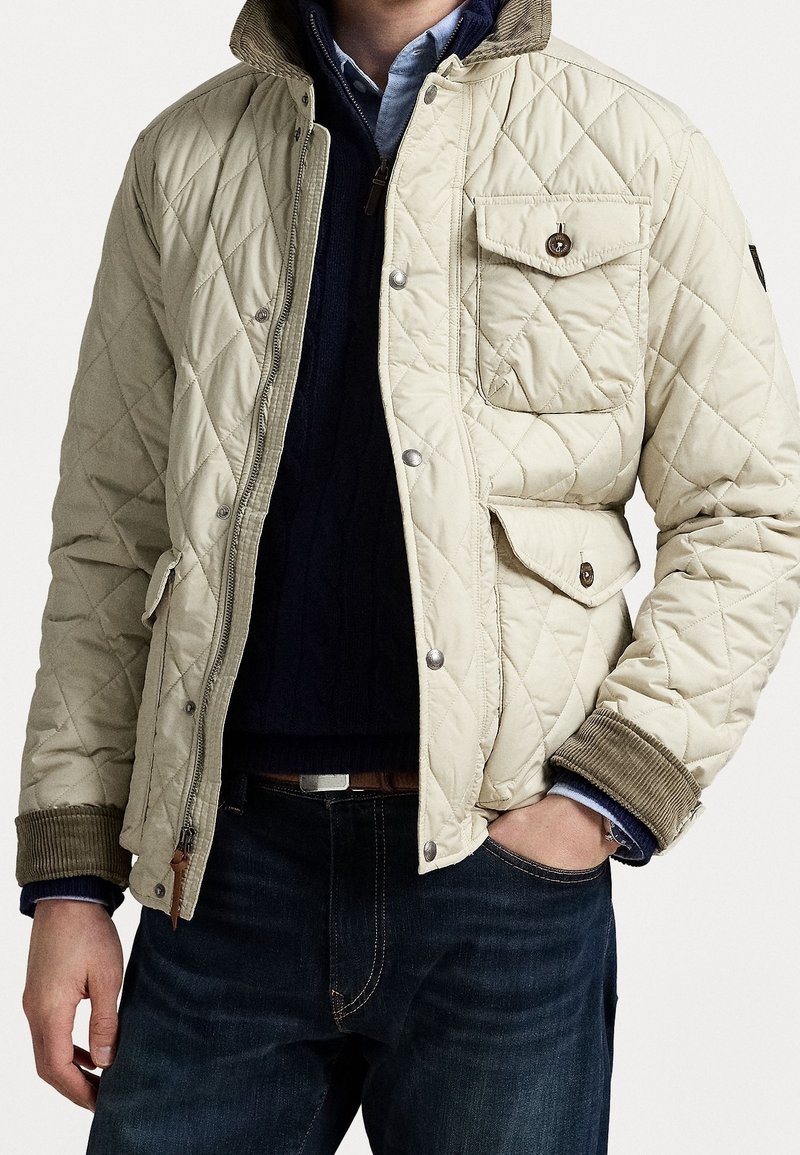 Quilted cream jacket with a corduroy collar, snap button pockets, and ribbed cuffs, worn over a dark sweater and denim jeans.