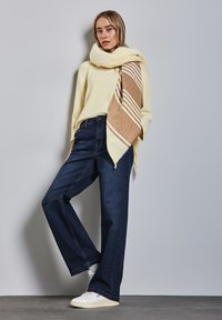 Suéter amarillo de gran tamaño de lana combinado con jeans de pierna ancha en azul oscuro y una bufanda beige y amarilla con patrón, llevado con zapatillas blancas.