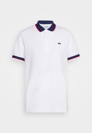 Polo blanca de algodón, con un collar azul marino con acentos rojos y un logo bordado verde en el pecho. Mangas cortas.