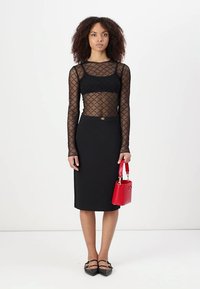 Blusa longa de malha preta com padrão de diamante, combinada com uma saia midi preta ajustada. Segura uma pequena mala vermelha e usa sapatos pretos planos.