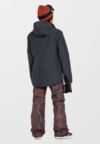 Schwarze Outdoor-Jacke mit Kapuze, kombiniert mit schnee- und braun-schwarzen Leopardenmuster-Hosen. Das Modell trägt eine rote Strickmütze und hält Handschuhe.