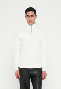 Mann in einem weißen Half-Zip-Pullover mit hohem Kragen und schwarzen Lederhosen, der vor einem schlichten weißen Hintergrund steht.