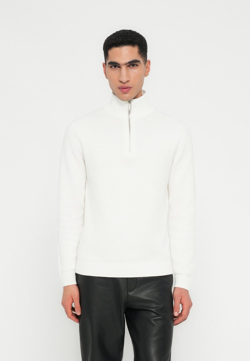 Mann in einem weißen Half-Zip-Pullover mit hohem Kragen und schwarzen Lederhosen, der vor einem schlichten weißen Hintergrund steht.