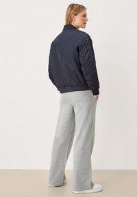 Vrouw die wegkijkt, gekleed in een marineblauwe bomberjack, lichtgrijze wijde broek, witte sneakers, staand tegen een effen lichte achtergrond.