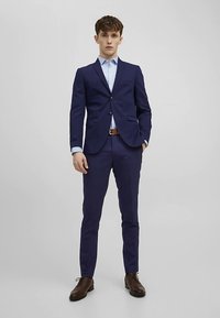 Tmavě modrý oblek s přiléhavým střihem, zahrnující jednobarevné sako a kalhoty. Pod ním bílá košile, hnědý pásek a boty.