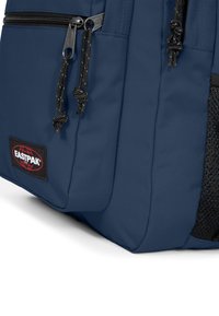 Mochila de tela azul que cuenta con compartimentos dobles con cremallera, bolsillo lateral de malla texturizada y un parche de marca negro cerca de la base.