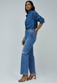 Jeans de denim azul claro con cintura alta y piernas rectas, que presentan detalles desgastados y cierre de cinco botones. Combinados con una blusa a juego.