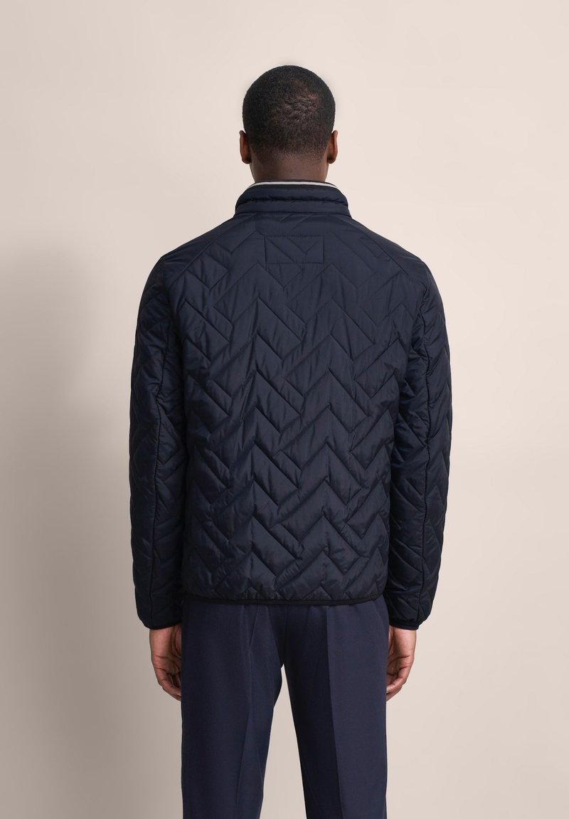 Dunkelblau gesteppte Jacke mit hohem Kragen, die ein Zickzackmuster und elastische Bündchen aufweist, getragen mit passenden dunklen Hosen.