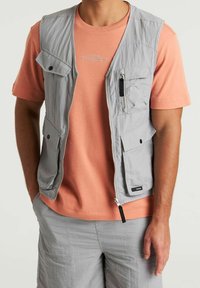 Gilet senza maniche grigio chiaro con molteplici tasche, cerniera frontale e tessuto testurizzato, indossato sopra una t-shirt color corallo. Design casual.