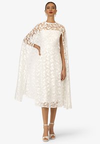 Witte kanten jurk met bloempatroon, doorzichtige cape, getailleerde bodice, knie-lange rok en hakken met enkelbandje. Glad materiaal met delicate details.