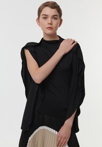 Top negro con capas, cuello alto y mangas drapeadas, combinado con una falda plisada negra y beis. Tejido suave con una silueta fluida.