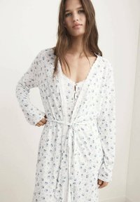 Robe blanche à motifs floraux bleus, manches longues et dentelle en finition. Entièrement nouée à la taille avec une ceinture assortie pour une silhouette définie.