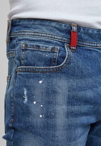 Primer plano de unos vaqueros de mezclilla azul con salpicaduras de pintura blanca, un pequeño parche desgastado, costuras naranjas y una etiqueta de marca roja cerca de la cintura.