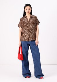 Kortärmad skjorta med leopardtryck och knäppning framtill, kombinerad med vida blå jeansbyxor. Modellen håller en liten röd väska.