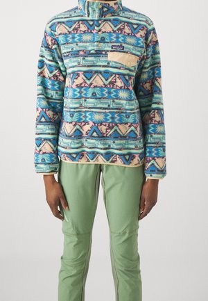 Personne portant un pull en polaire à motifs bleu, rose et vert avec une poche poitrine beige et un pantalon vert clair sur un fond neutre.