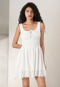 White sleeveless dress with a square neckline, smocked waist, and embroidered eyelet detailing at the hem. Soft cotton fabric, A-line shape.