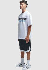 Tricou alb supradimensionat cu text grafic, asociat cu pantaloni scurți atletici negri cu accente albe și adidași albi cu dungi negre.