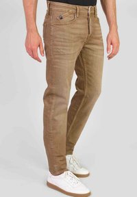 Bruine spijkerbroeken met een slim fit, voorzien van gestikte accenten en voorzakken. Geshowd met witte sneakers op een lichtgrijze achtergrond.