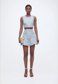 Γυναίκα που φοράει ανοιχτό μπλε αμάνικο crop top και ταιριαστό μίνι φούστα με βολάν, κρατά ένα μικρό μπεζ τσαντάκι χειρός και φοράει μπεζ ψηλοτάκουνα.