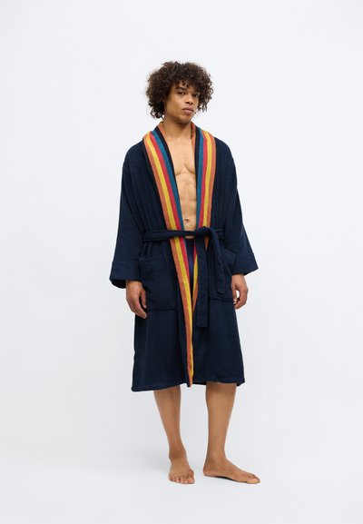 Noor mees, kes seisab paljajalu, kandes tumesinist hommikumantlit, mille krae ääres on oranžid, punased, kollased ja sinised triibud, valge taustaga.