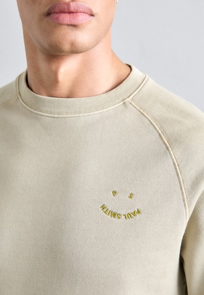 Béžová mikina s žebrovaným výstřihem a švy. Obsahuje žlutý vyšívaný úsměv s textem. Měkká tkanina, ležérní design.