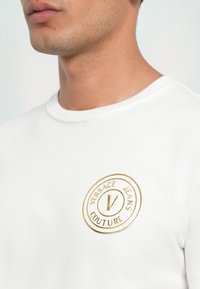 Hvit bomulls t-skjorte med et rundt, gullbrodert logo med teksten "VERSACE JEANS COUTURE" og sentralt "V"-design. Klassisk rund nakkekrage.