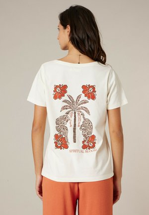 Femme portant un t-shirt blanc avec un imprimé au dos représentant un palmier, deux léopards, des fleurs orange, et le texte "SPIRITUAL BLOOM".
