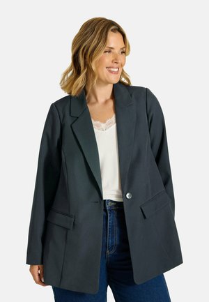 Donna con capelli biondi fino alle spalle indossa un blazer grigio scuro sopra una maglia bianca con bordi in pizzo e jeans blu, sorride e guarda verso destra.