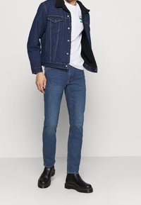 Denim jacket with black shearling collar, white t-shirt featuring a cactus graphic, blue slim-fit jeans, and black chelsea boots with yellow stitching.