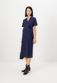 Σκούρο μπλε μαιευτική φόρμα με κοντά μανίκια, κουμπάκι μπροστά και ζώνη στη μέση. Διαθέτει πλευρικές σχισμές και λεία υφή. Φοράει μαύρες μπότες.