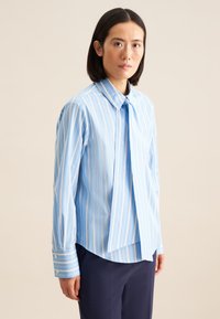 Camicia a righe blu chiaro con colletto button-down e cravatta lunga. Presenta righe verticali bianche e dettagli a contrasto sui polsini.