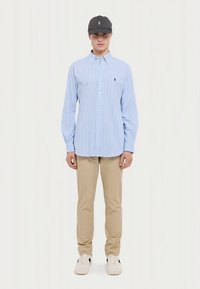 Gaiši zila svītraina pogājama krekla, kaki krāsas bikses un pelēka cepure ar logo. Modelis valkā baltas sporta kurpes. Formāls, bet vienlaikus ikdienas apģērbs.