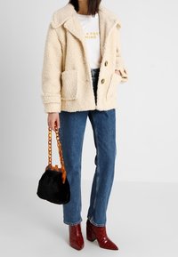 Krémová fleecová bunda s velkým límcem a předními kapsami, ve spojení s modrými rovnými džíny, červenými lodičkami na vysokém podpatku a černou kožušinovou kabelkou s řetízkovým popruhem.