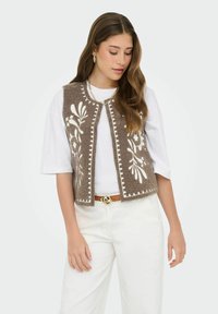 Jeune femme aux longs cheveux portant un gilet brodé de fleurs marron sur une chemise blanche, un pantalon blanc, des bijoux dorés et une ceinture marron.