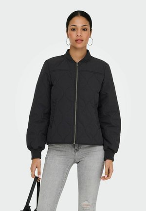 Woman wearing black quilted zip-up jacket, light gray ripped jeans, large hoop earrings, holding black handbag strap, standing against white background.