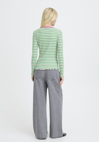 Eine Frau mit langen blonden Haaren trägt ein grün-weiß gestreiftes Langarmoberteil und lockere graue Jeans, steht vor einem weißen Hintergrund, Rückansicht.