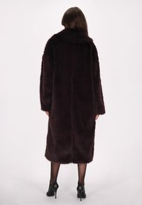 Lang, mørk burgunder faux fur frakke med bred krave, oversized pasform og blød tekstur, der når til midten af læggen, parret med sorte stiletto hæle.