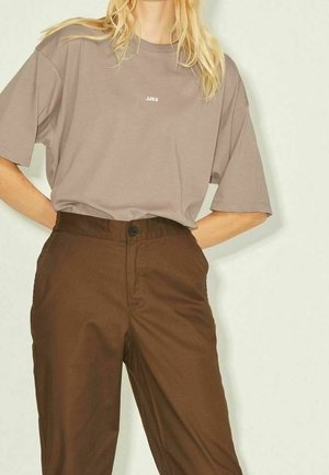 Femme portant un t-shirt ample taupe avec le texte « JJXX » et un pantalon marron taille haute, debout les mains derrière le dos devant un fond uni.