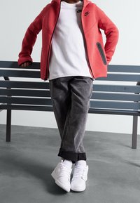 Czerwona bluza z zamkiem błyskawicznym z czarnymi akcentami, biała koszulka, szare dżinsy, i białe sneakersy typu high-top, stojąca na szarym podłożu.