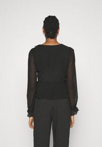 Blusa nera trasparente a maniche lunghe con scollatura rotonda, dettagli arricciati sulle spalle e polsini elastici; abbinata a pantaloni a gamba larga a righe.