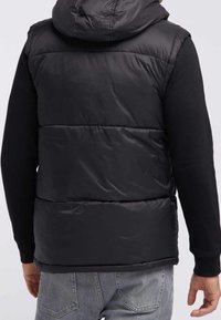 Chaleco acolchado negro con capucha, que presenta un diseño acolchado, textura volumétrica y una construcción sólida. Llevado sobre una camiseta negra de manga larga.