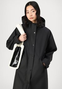 Svart vattentät kappa med huva, med tryckknappar, sidofickor och en vit pälskonstruktion strap-väska med insats i svart nät.