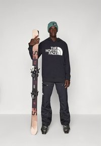 Camisola com capuz preta da The North Face, calças de esqui azul-marinho, gorro verde. Segurando um esqui com fixações, madeira clara com detalhes em rosa escuro.